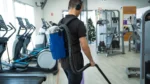 Man wearing headphones and carrying a blue backpack-style vacuum walks through a modern gym filled with cardio