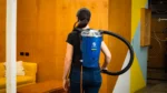 Woman wearing a blue backpack vacuum cleaner in an orange-walled room.
